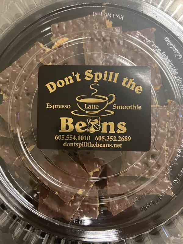 Clear plastic container labeled 'Don't Spill the Beans' containing Scotcharoo Bars, 24 precut and ready to serve. Contact info and website are included on the label.