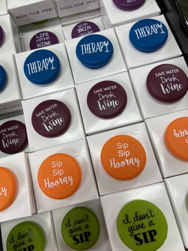 CapaBunga offers a variety of wine bottle stoppers featuring colorful designs and humorous text, including phrases like "Save Water, Drink Wine" and "Sip Sip Hooray.