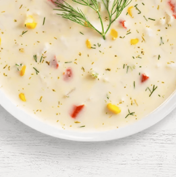 A bowl of Florida Sunshine Red Pepper Corn Chowder with a creamy texture, showcasing visible corn, red bell pepper pieces, herbs, and a green garnish on top, set on a white wooden surface.
