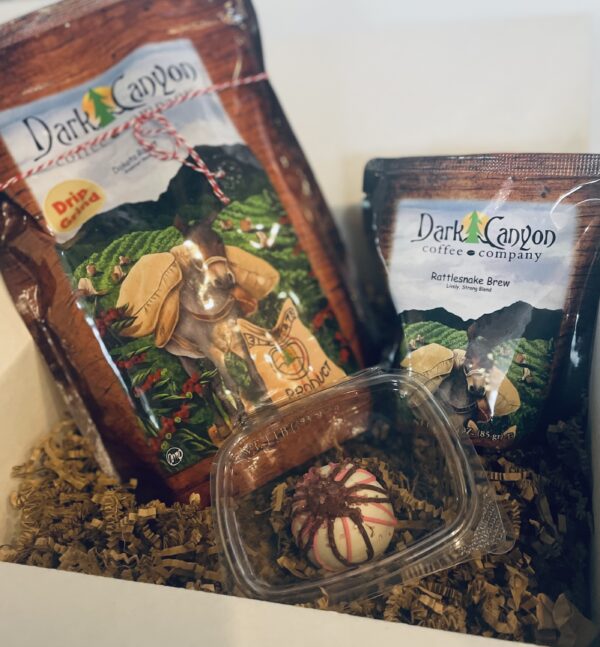 A Coffee Gift Box & Subscription featuring a large coffee bean bag, a smaller "Dark Canyon" coffee packet, and a decorative chocolate truffle in a clear container nestled on shredded packing material.