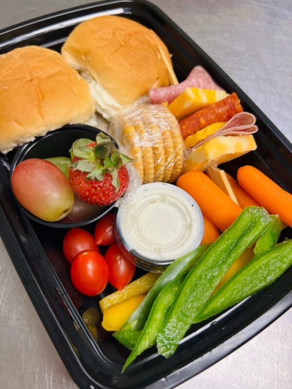 Introducing The Cornerpost Grapefarm Charcuterie Box: a black tray featuring two bread rolls, an assortment of meats, cheese slices, crackers, grapes, strawberries, cherry tomatoes, carrots, bell pepper slices, and a delicious dipping sauce.