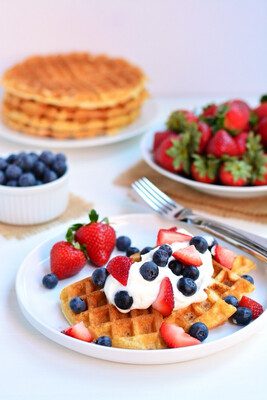 A waffle adorned with whipped cream, blueberries, and strawberries sits on a plate. In the background, there's a bowl of blueberries, a plate of strawberries, and an additional stack of Waffles. A fork and knife are placed nearby.