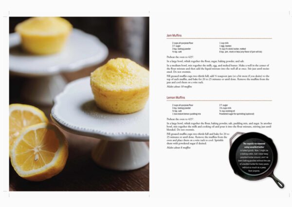 Open the Homestead To Table Cookbook featuring recipes for jam muffins and lemon muffins, placed next to two baked muffins on a white stand with sliced lemons.
