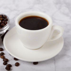 A cup of House Brewed Coffee in a white mug on a saucer rests on marble, surrounded by scattered coffee beans and a small glass bowl filled with them.