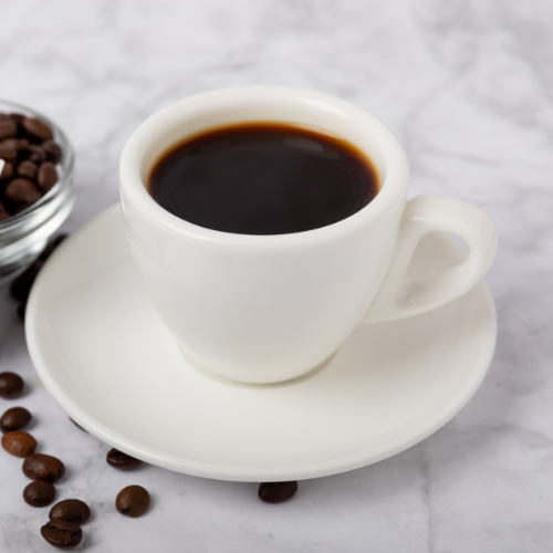 A cup of House Brewed Coffee in a white mug on a saucer rests on marble, surrounded by scattered coffee beans and a small glass bowl filled with them.