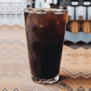 A glass of Iced House Brewed Coffee sits on a wooden table with a blurred background.