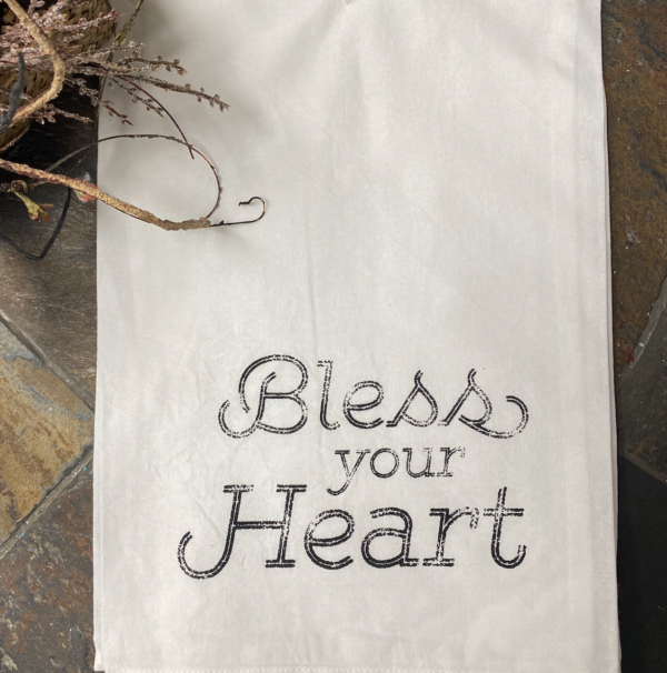A white cloth with the words "Bless your Heart" printed in black on a tiled surface, with a branch of dried flowers in the top left corner.