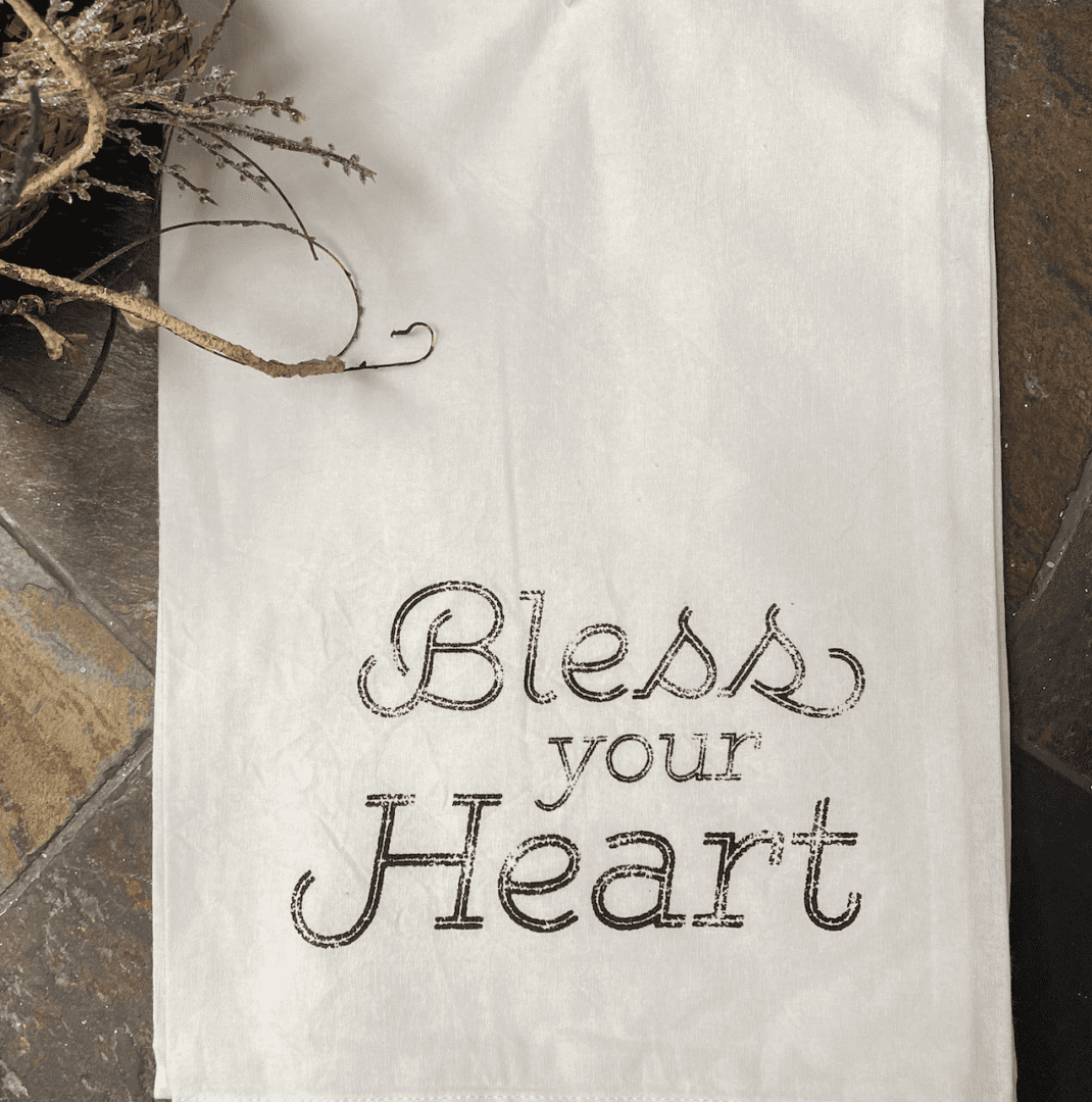 A white cloth with the words "Bless your Heart" printed in black on a tiled surface, with a branch of dried flowers in the top left corner.