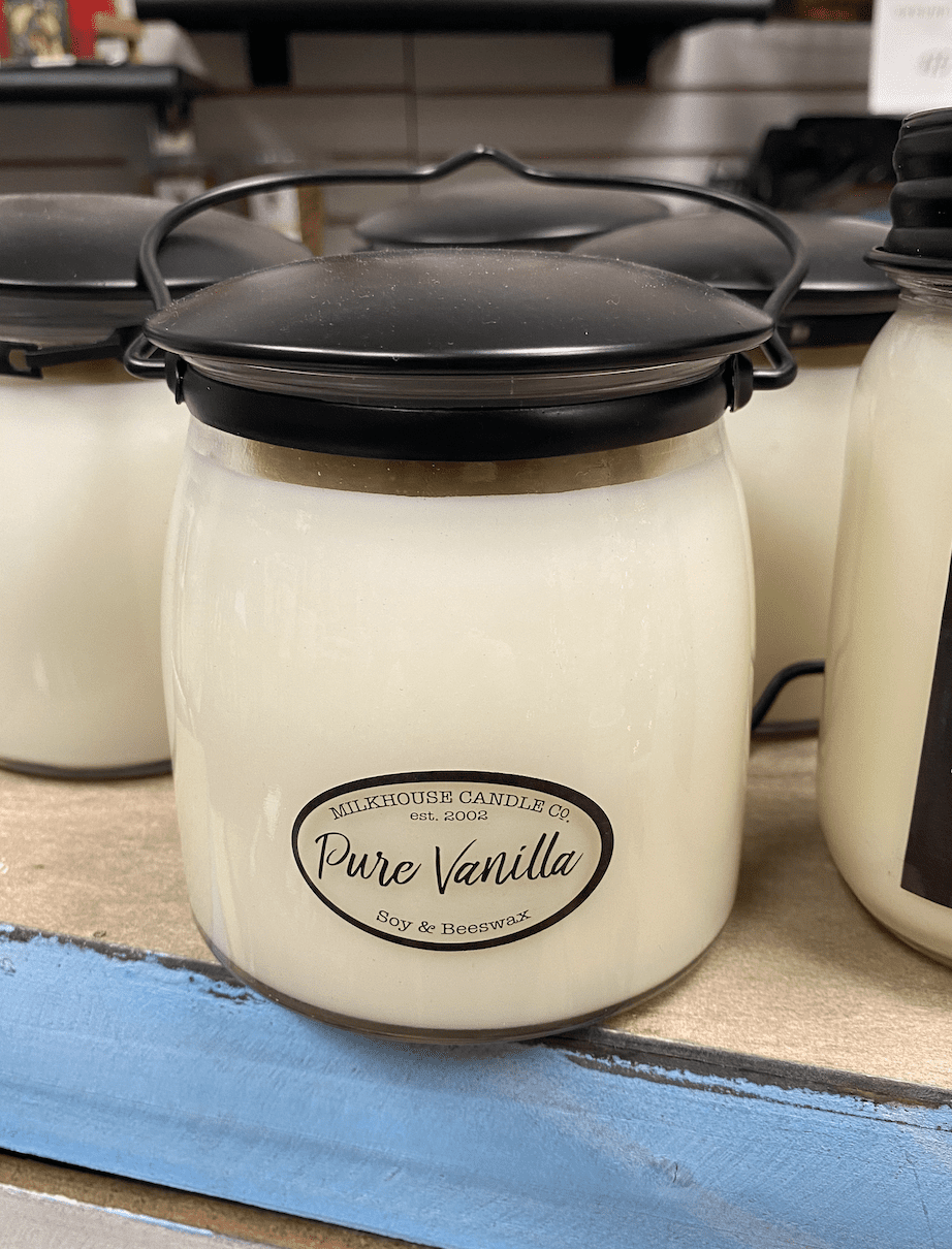 A white soy and beeswax candle in a glass jar with a black lid, labeled "Pure Vanilla" from Milkhouse Candle Co., on a shelf.