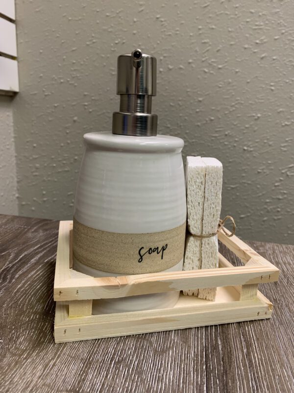 Ceramic soap dispenser and sponge on a wooden tray, placed on a textured surface against a speckled wall.