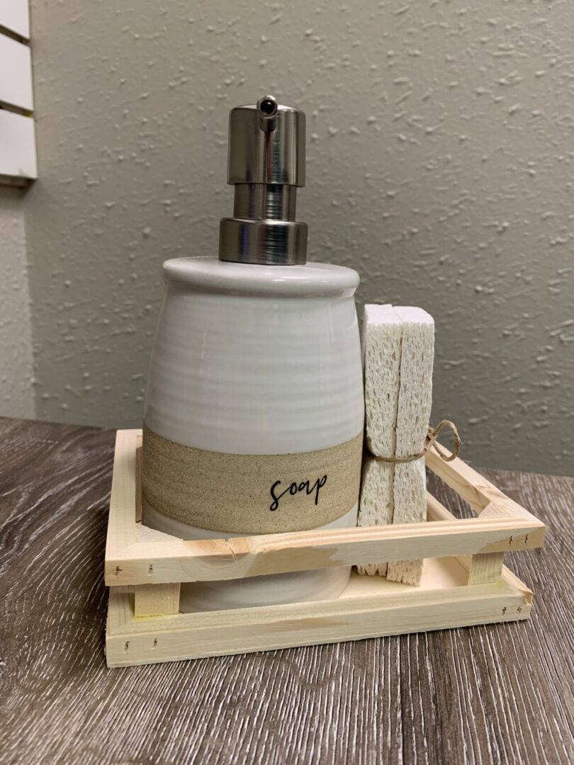 Ceramic soap dispenser and sponge on a wooden tray, placed on a textured surface against a speckled wall.