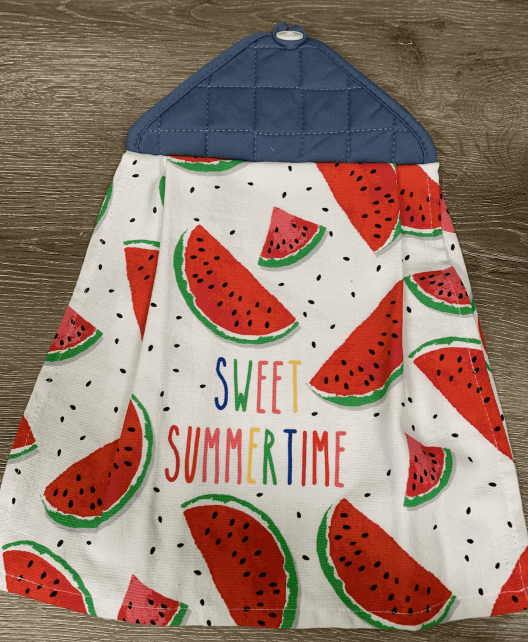 A kitchen towel featuring a watermelon pattern with the words "Sweet Summertime" in colorful letters, hanging on a wood surface.