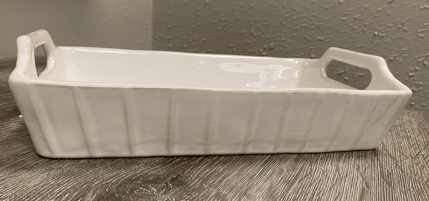 White rectangular ceramic baking dish with handles on a wooden surface against a textured wall.