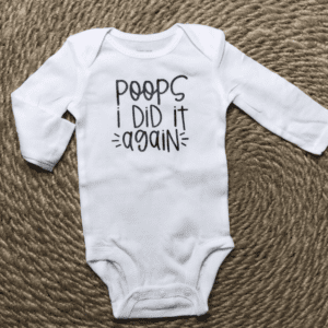 White baby onesie on a woven rug, with the text "Poops I did it again" printed on the front in black letters.