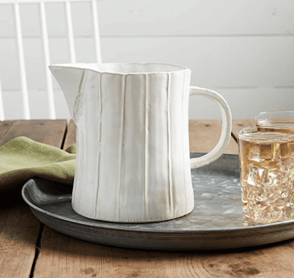 A textured white ceramic pitcher and two small glasses sit on a round metal tray on a wooden table. A folded green napkin is nearby.