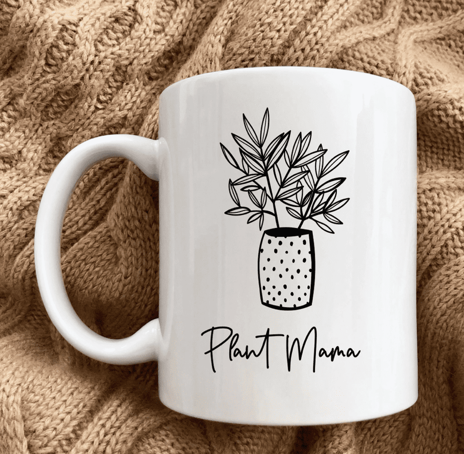 White mug with black line art of a potted plant and the text "Plant Mama," placed on a textured beige fabric.