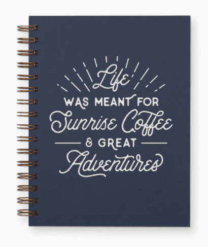 A navy blue spiral notebook with the words "Life was meant for sunrise coffee & great adventures" printed in white.
