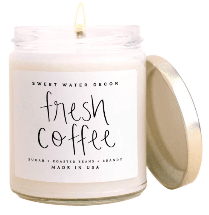 A lit candle in a clear jar with a gold lid. The label reads "Sweet Water Decor, fresh coffee, sugar, roasted beans, brandy, Made in USA.