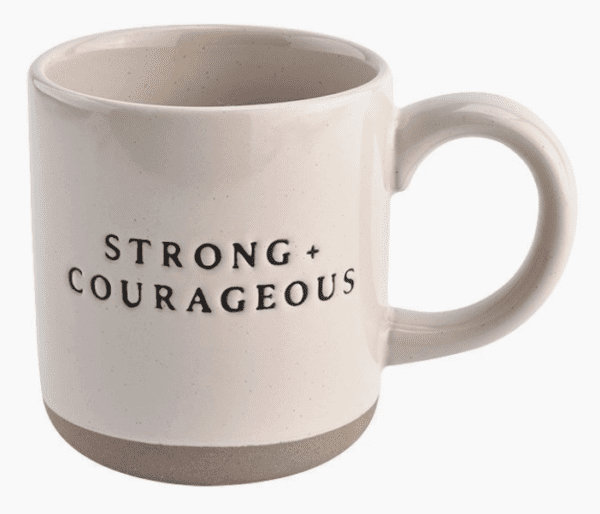 A white ceramic mug with a large handle, featuring the text "STRONG + COURAGEOUS" in black letters.