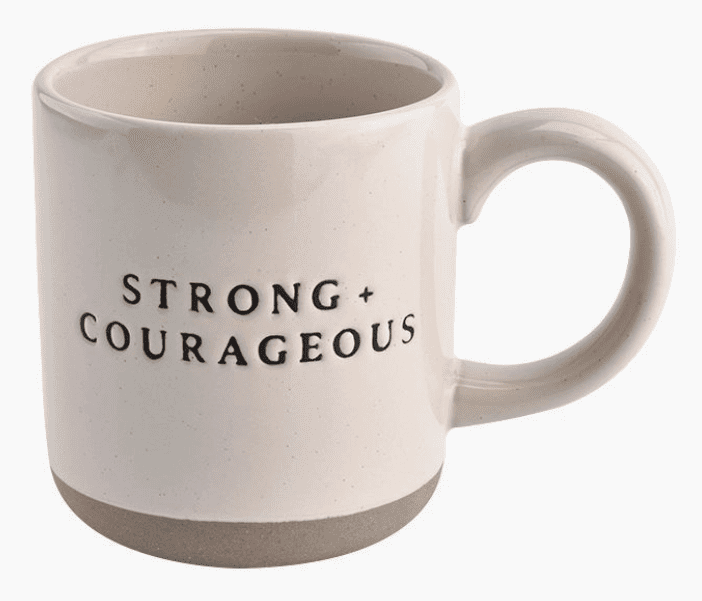 A white ceramic mug with a large handle, featuring the text "STRONG + COURAGEOUS" in black letters.