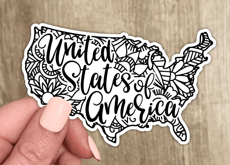 A hand holds a decorative sticker shaped like the United States with "United States of America" written in cursive, featuring floral patterns.
