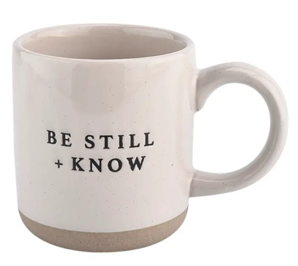 A white mug with "Be Still + Know" printed in black text on its side.