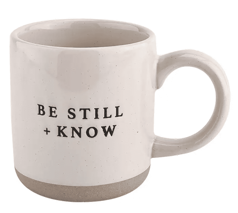 A white mug with "Be Still + Know" printed in black text on its side.