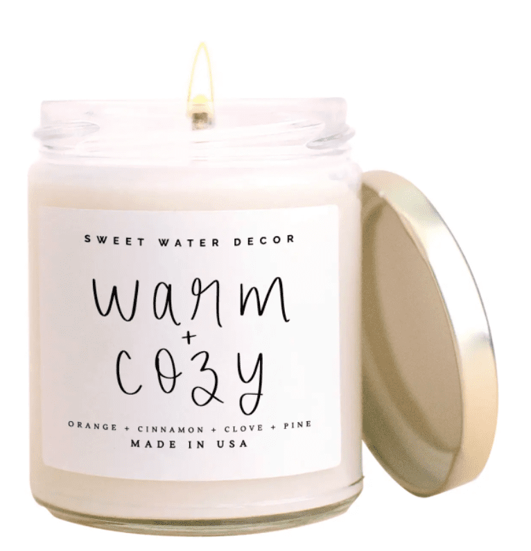 A lit candle in a jar with a gold lid, labeled "Warm + Cozy." Scents listed: orange, cinnamon, clove, and pine. Made in USA.