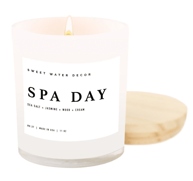White candle labeled "SPA DAY" with scents of sea salt, jasmine, wood, and cream. The candle is lit and placed beside a wooden lid. Made in the USA, 11 oz.