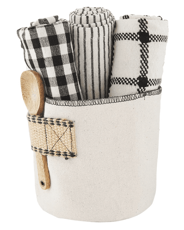 A cream fabric storage basket holds three rolled black and white patterned dish towels and a wooden spoon in a side loop.