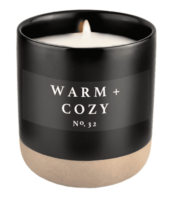A black candle labeled "Warm + Cozy No. 32" with a lit wick, in a matte black container with a beige base.