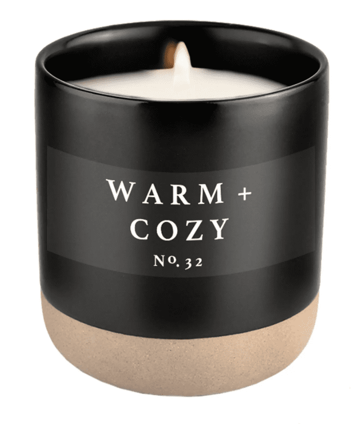 A black candle labeled "Warm + Cozy No. 32" with a lit wick, in a matte black container with a beige base.