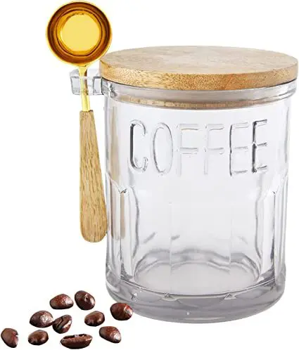 A glass jar labeled "coffee" with a wooden lid and a golden coffee scoop attached. Coffee beans are scattered beside it.