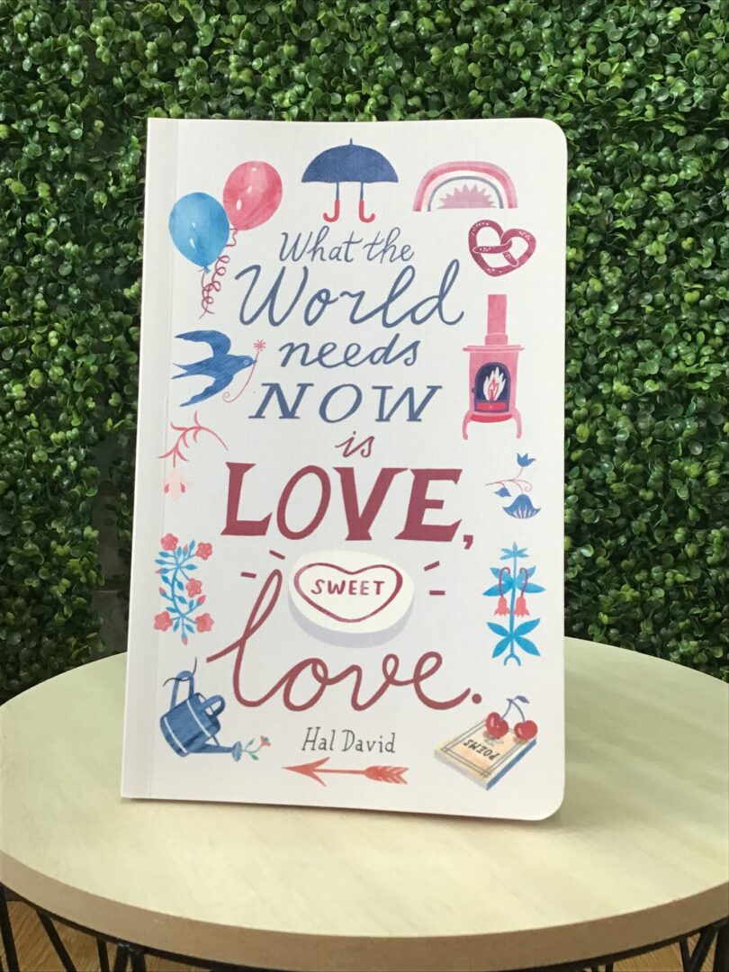 A book titled "What the World Needs Now is Love, Sweet Love" by Hal David, featuring illustrations of balloons, flowers, and hearts, is displayed on a wooden table with a leafy background.
