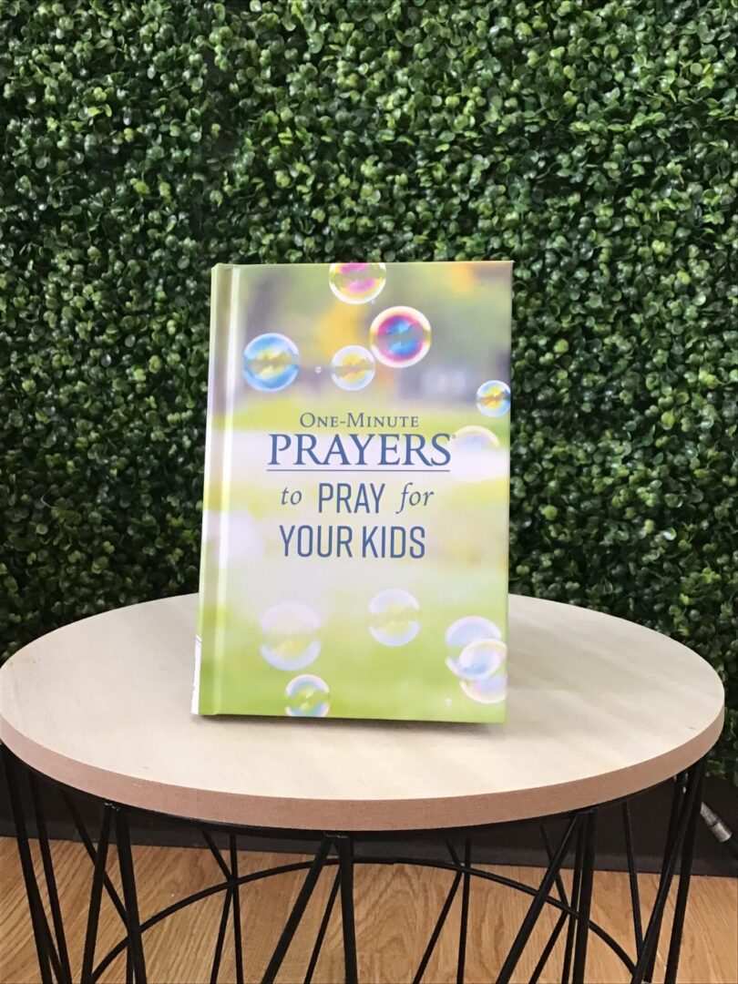 A book titled "One-Minute Prayers to Pray for Your Kids" stands on a round table against a green leafy background.