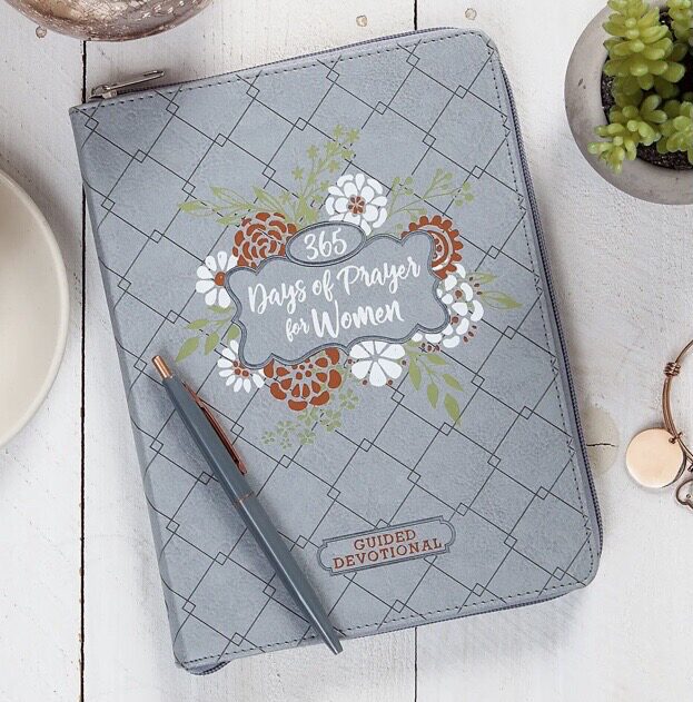 A gray zippered devotional book titled "365 Days of Prayer for Women" lies on a white table, accompanied by a pen and a small potted plant.