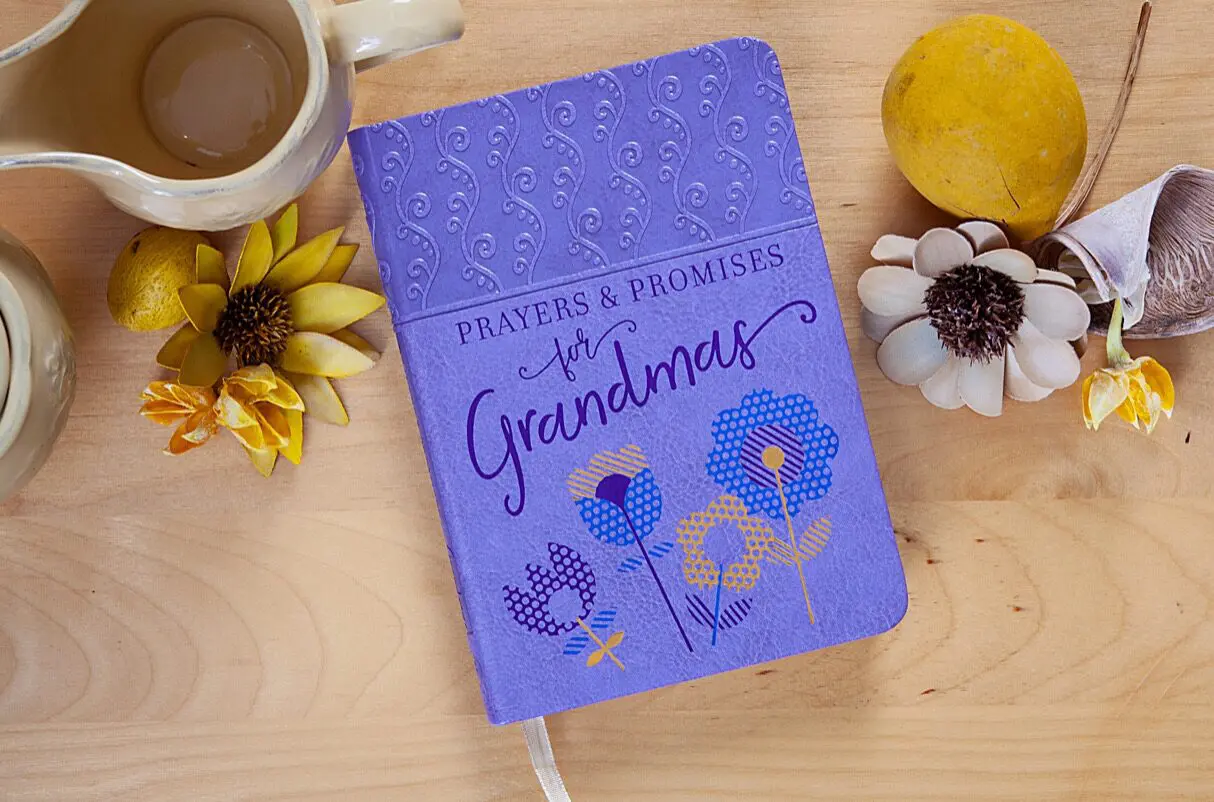 Purple book titled "Prayers & Promises for Grandmas" on a wooden table with flowers, a yellow fruit, and a ceramic pitcher.