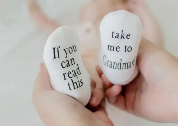 Baby wearing socks with "If you can read this" and "take me to Grandma's!" text, held in adult hands.