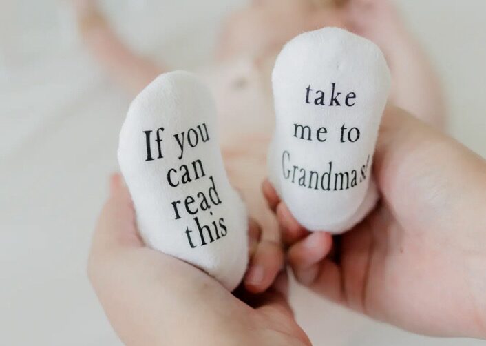 Baby wearing socks with "If you can read this" and "take me to Grandma's!" text, held in adult hands.