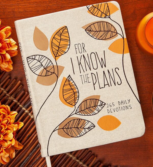 A journal titled "For I Know The Plans: 365 Daily Devotions" with leaf illustrations, surrounded by orange flowers and a wooden mat.