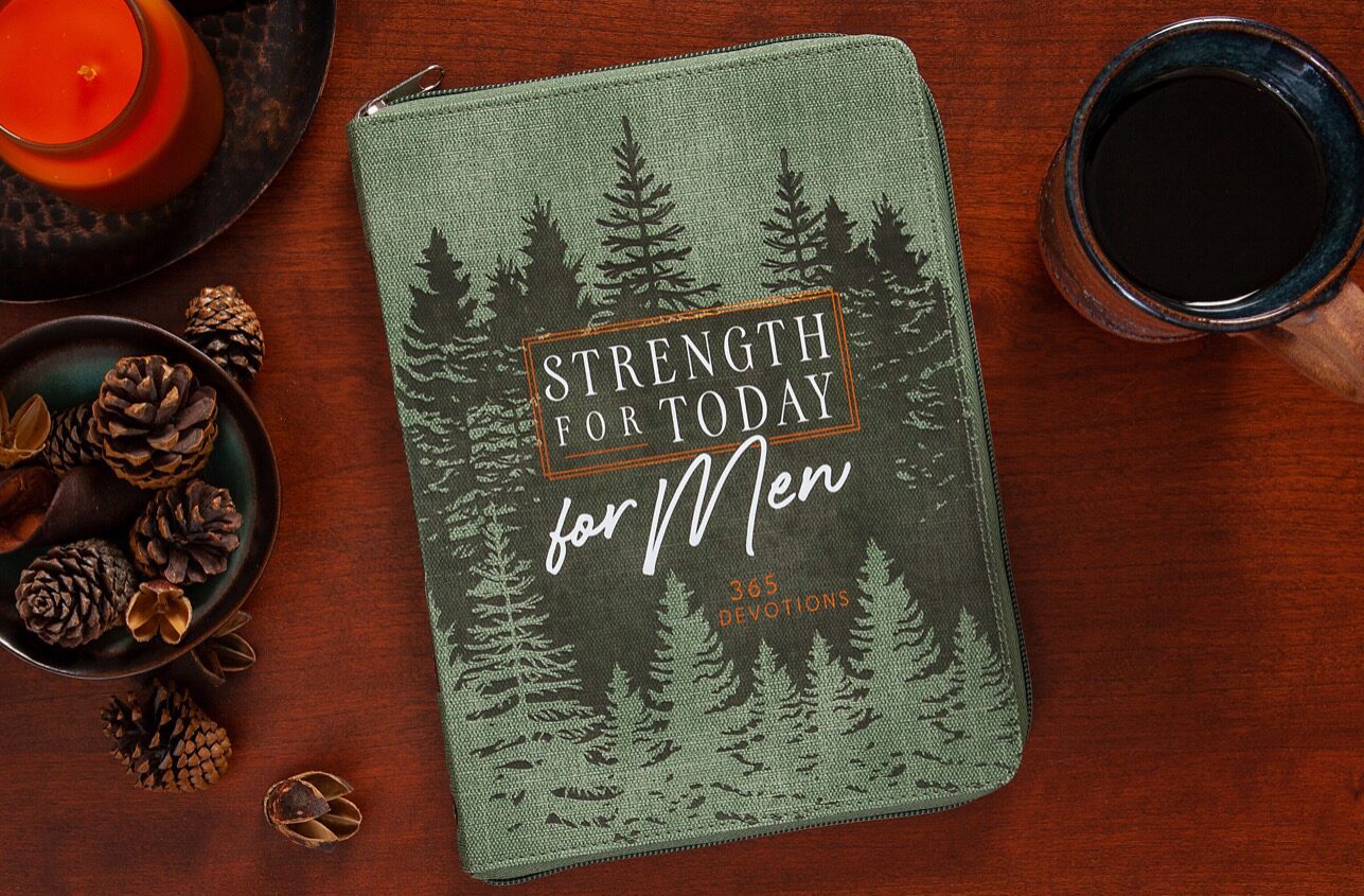 Book cover with "Strength for Today for Men: 365 Devotions" on a forest design background. Nearby are a candle, pinecones, and a mug on a wooden table.