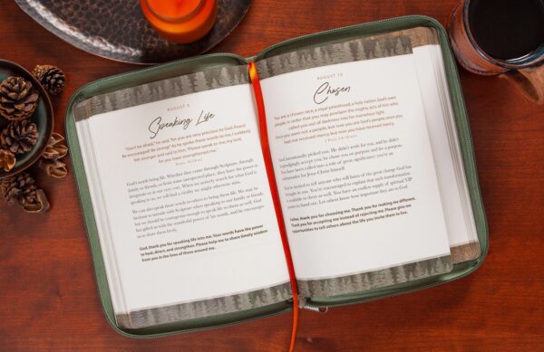 The "Strength For Today Men’s Devotional" book is open on a wooden table, accompanied by pinecones, a candle, and a cup. The pages are dated August 9 and 10, with a red ribbon bookmark draped across the center.