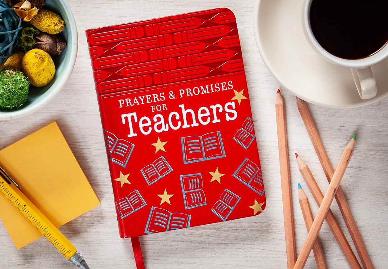 Red notebook titled "Prayers & Promises for Teachers" on a table with colored pencils, a coffee cup, sticky notes, and a plant.