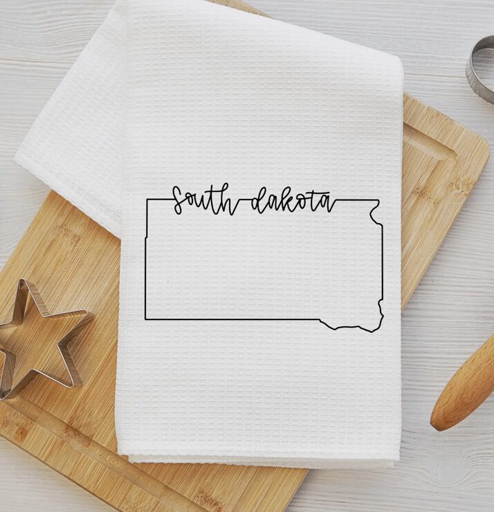 A kitchen towel with the outline of South Dakota and text "South Dakota" placed on a wooden cutting board with a star-shaped cookie cutter and rolling pin nearby.