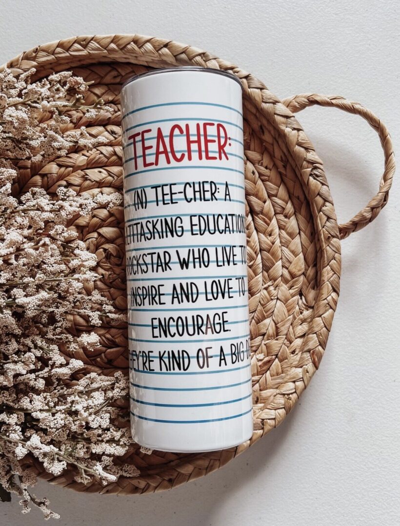 A tall mug with text defining "Teacher" as a multitasking educator. It rests on a woven tray next to dried flowers.
