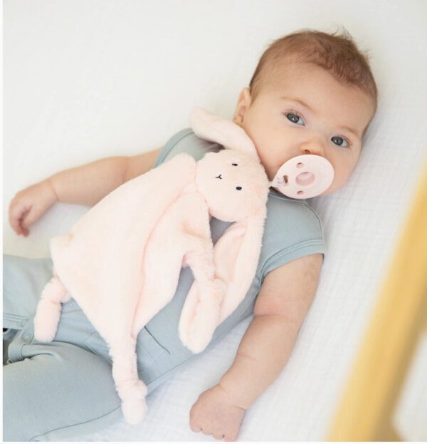 A baby is lying down with a pacifier in their mouth, holding the Bunny Bubbi Buddy, a soft pink bunny-shaped blanket, while wearing a light blue outfit.