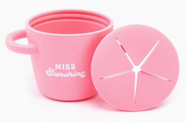A pink Miss Sunshine Snacker with "Bella Tunno" and "Miss Sunshine" printed on it, featuring a removable lid with a star-shaped opening.