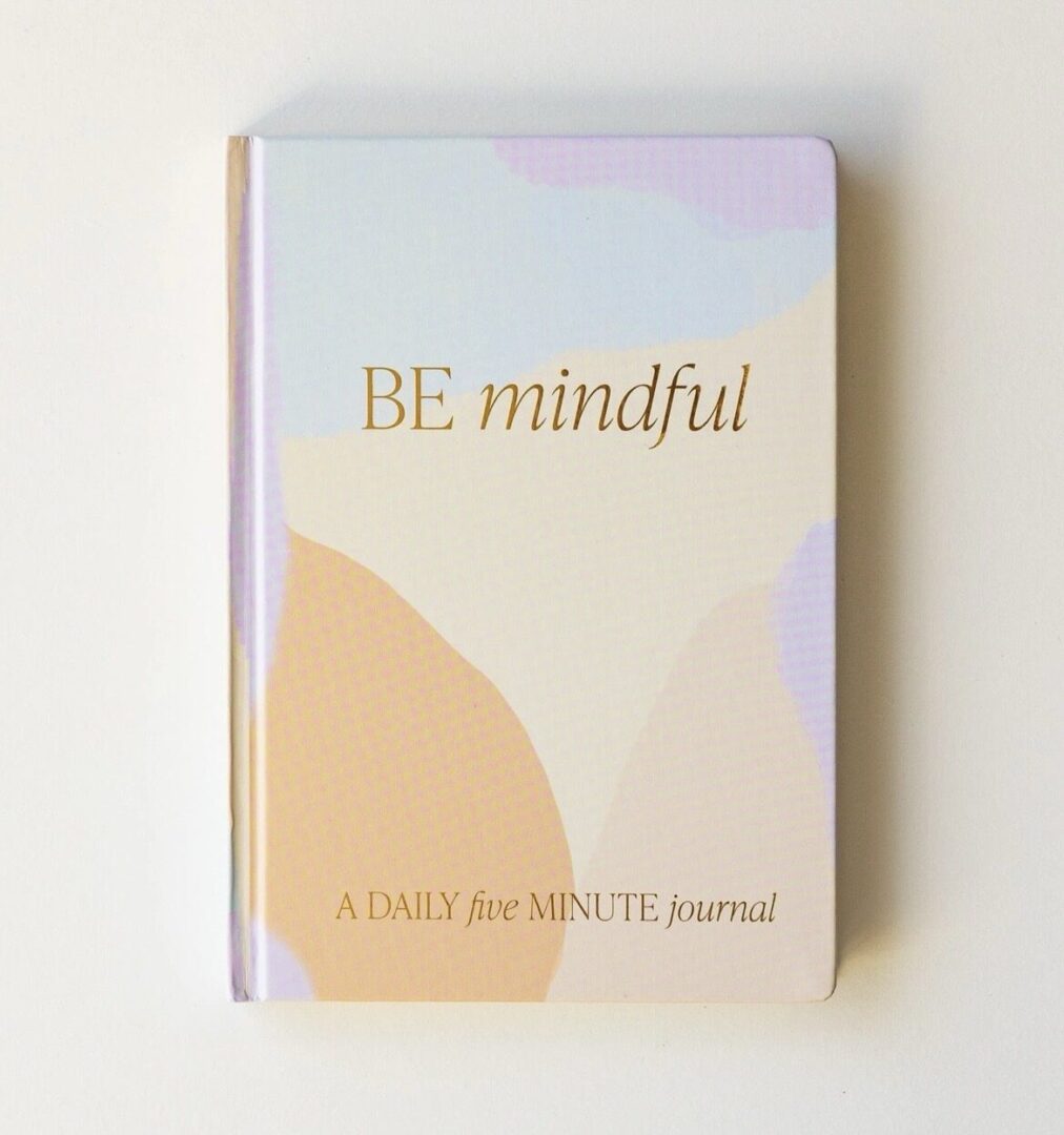 A journal with a pastel abstract cover features the text "BE mindful" and "A DAILY five MINUTE journal.