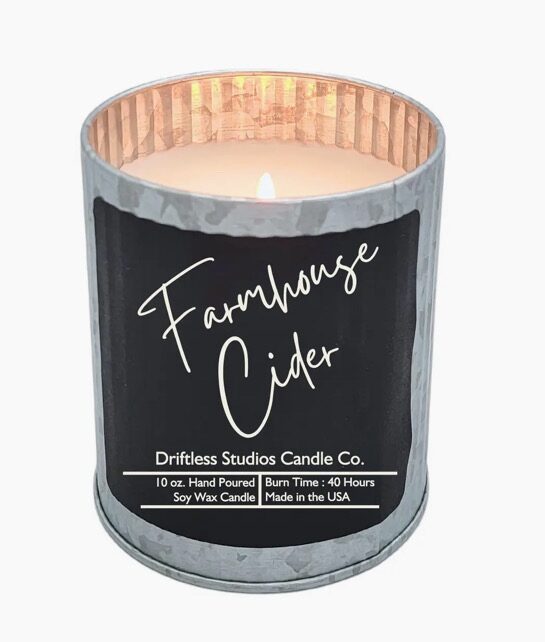 A lit candle in a metal container labeled "Farmhouse Cider" from Driftless Studios Candle Co., with details about size, burn time, and origin.