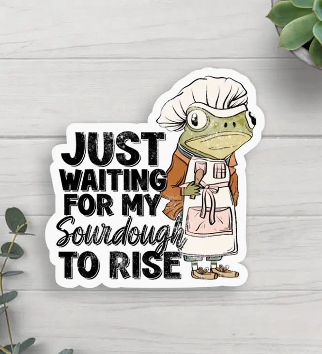 Cartoon frog in chef's outfit holding an apron with text: "Just waiting for my sourdough to rise." Background has plants on a white wooden surface.